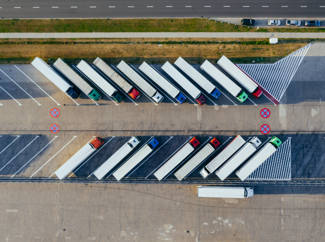 Aerial view of a parking lot with a row of mostly white trucks parked in designated spaces, adjacent to a road with passing cars, and pavement markings including a no-entry sign.