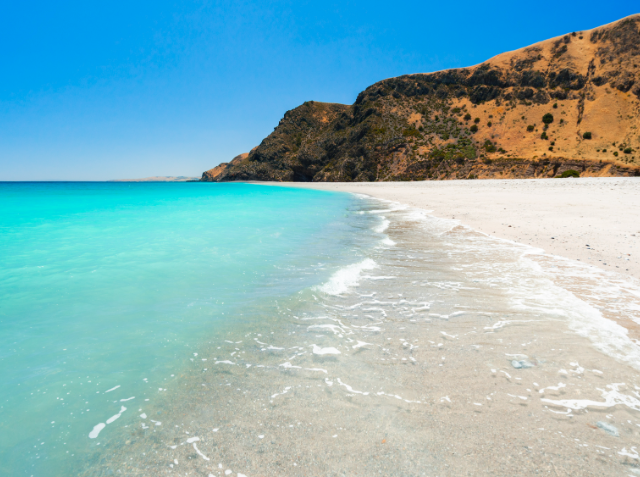 Serene beach with white sand and clear turquoise waters, gentle waves at the shore, and a rugged hill with brownish vegetation under a clear blue sky.