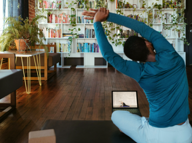 Person practicing yoga in a room with a laptop open in front of them, surrounded by bookshelves and indoor plants.