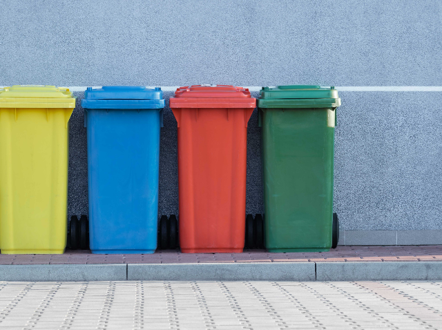 Four colored wheelie bins in yellow, blue, red, and green lined up against a light grey wall with a white horizontal line above them, on a paved surface with tactile paving in the foreground.