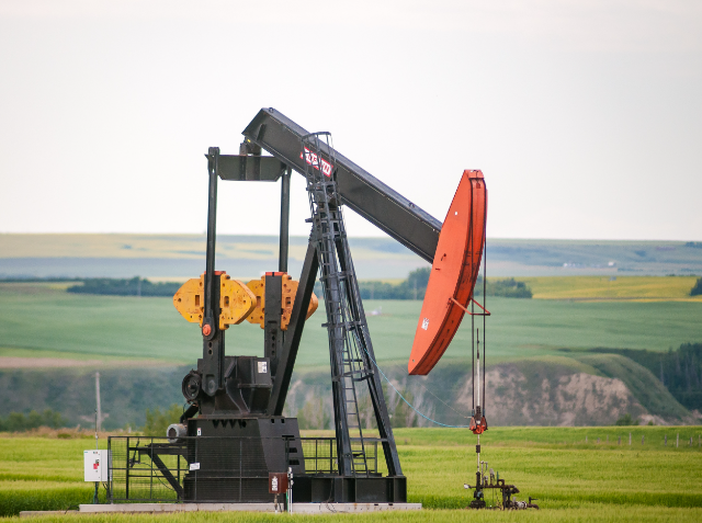 A black and orange pumpjack operates in a green field with rolling hills in the background.