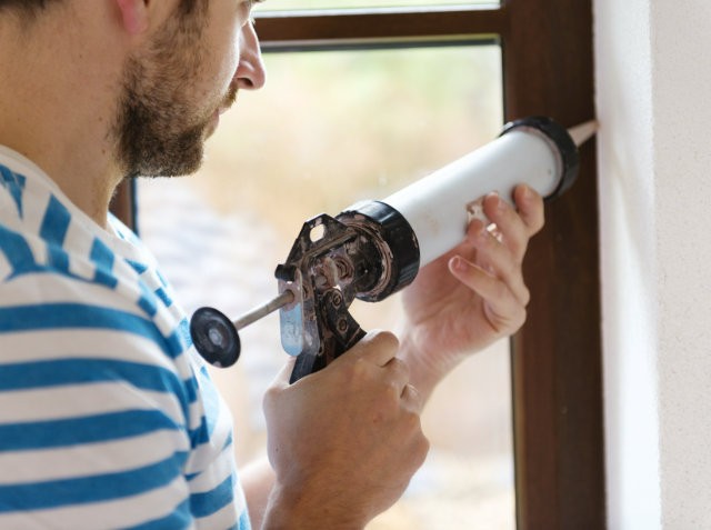 Person applying caulk to a window frame with a caulking gun.