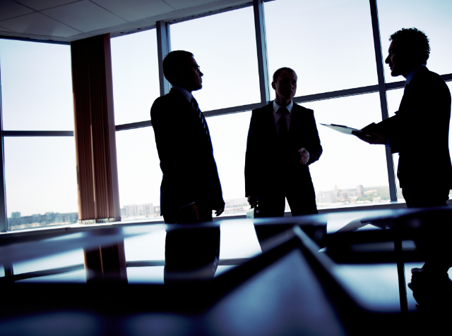 Silhouetted image of three businessmen in a discussion inside an office with large windows overlooking a city skyline.