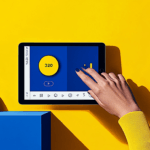 A hand with blue nail polish uses a tablet displaying the number 320, on a yellow and blue background with a yellow ball.