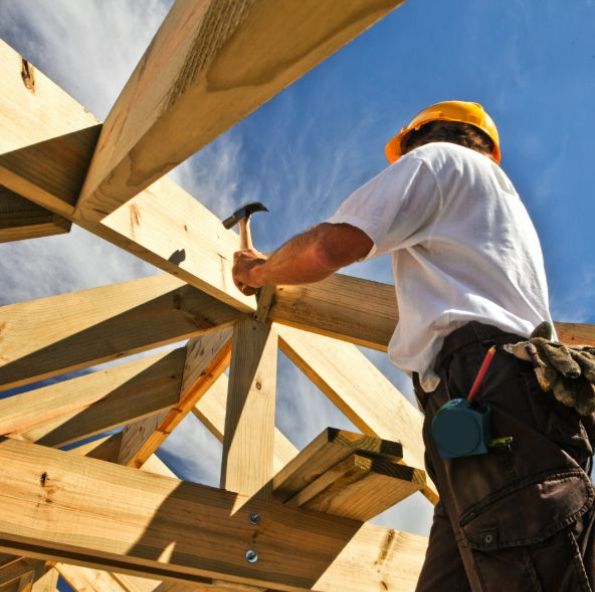 A construction worker in a hard hat hammers wooden beams against a bright blue sky, focusing on the structure above