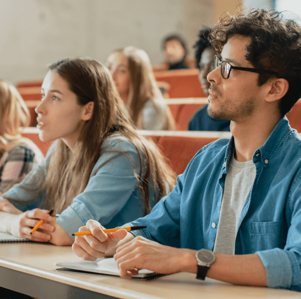 Students taking notes in tiered lecture hall, focused young man in blue shirt and glasses in foreground