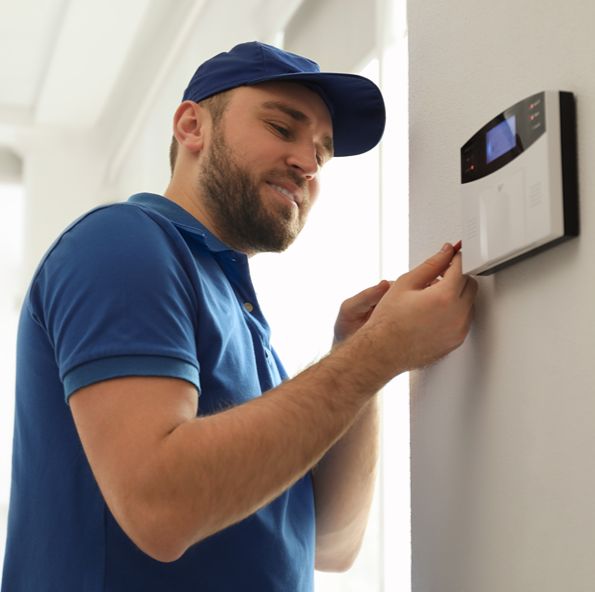 A technician in a blue cap and polo smiles while adjusting a wall-mounted alarm keypad