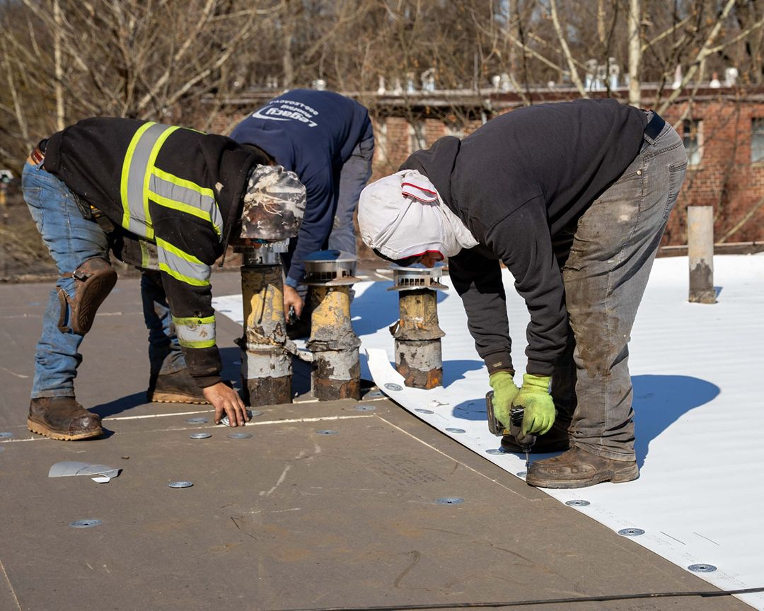 Crew fastening white TPO roofing membrane around vent pipes